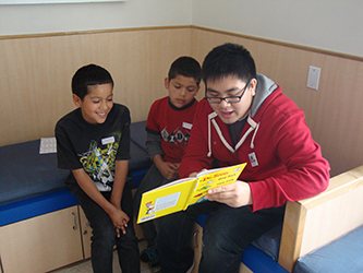 A teen volunteer reading a picture book to two younger children in a TCC clinic exam room